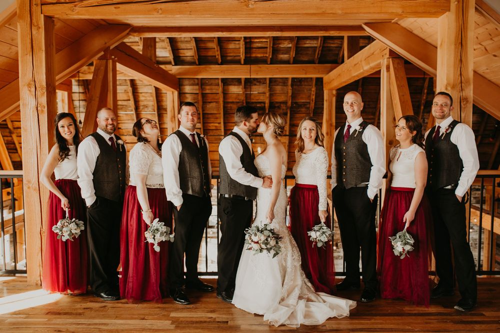 Wedding party poses in rustic barn venue, bridesmaids in burgundy gowns flanking bride and groom.