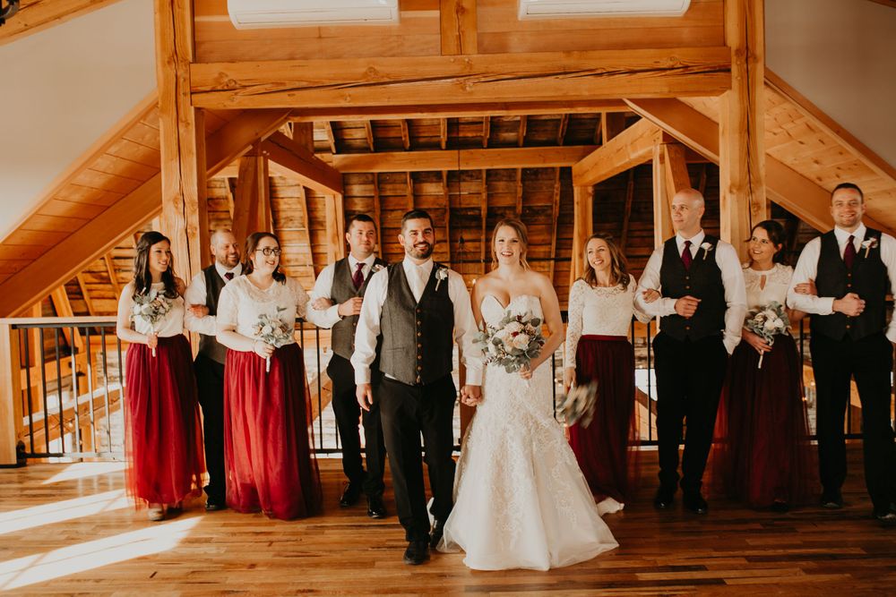 Couple leads wedding party walk in sunlit barn, bridesmaids in burgundy and groomsmen in dark suits.