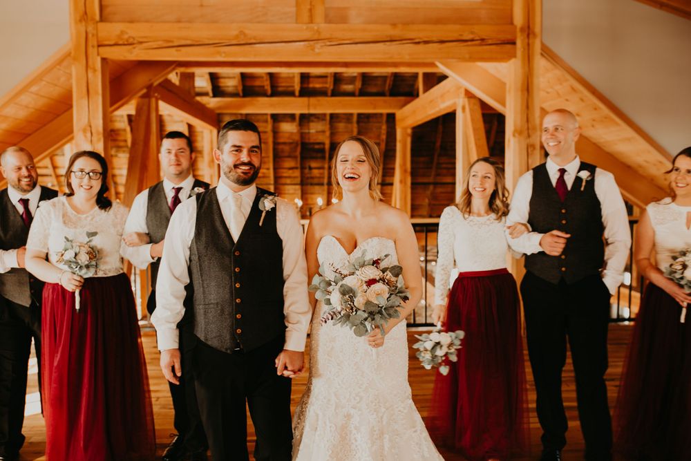 Laughing bride and groom hold hands surrounded by joyful wedding party in rustic barn venue.
