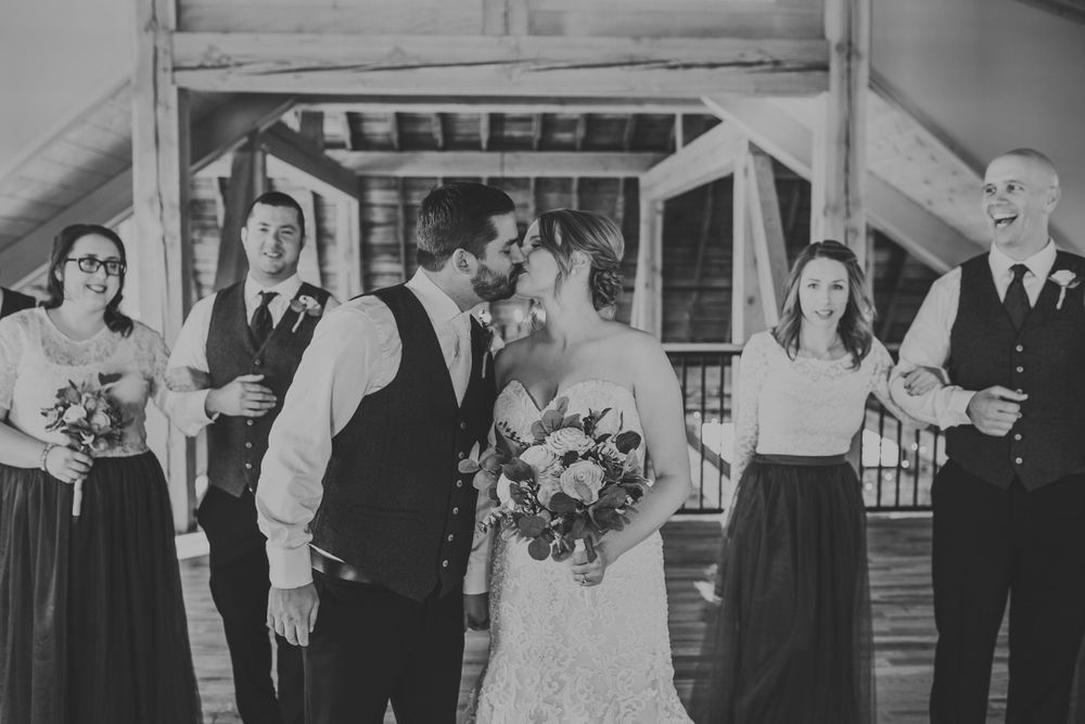 Black and white photo of bride and groom sharing a kiss with wedding party celebrating around them.