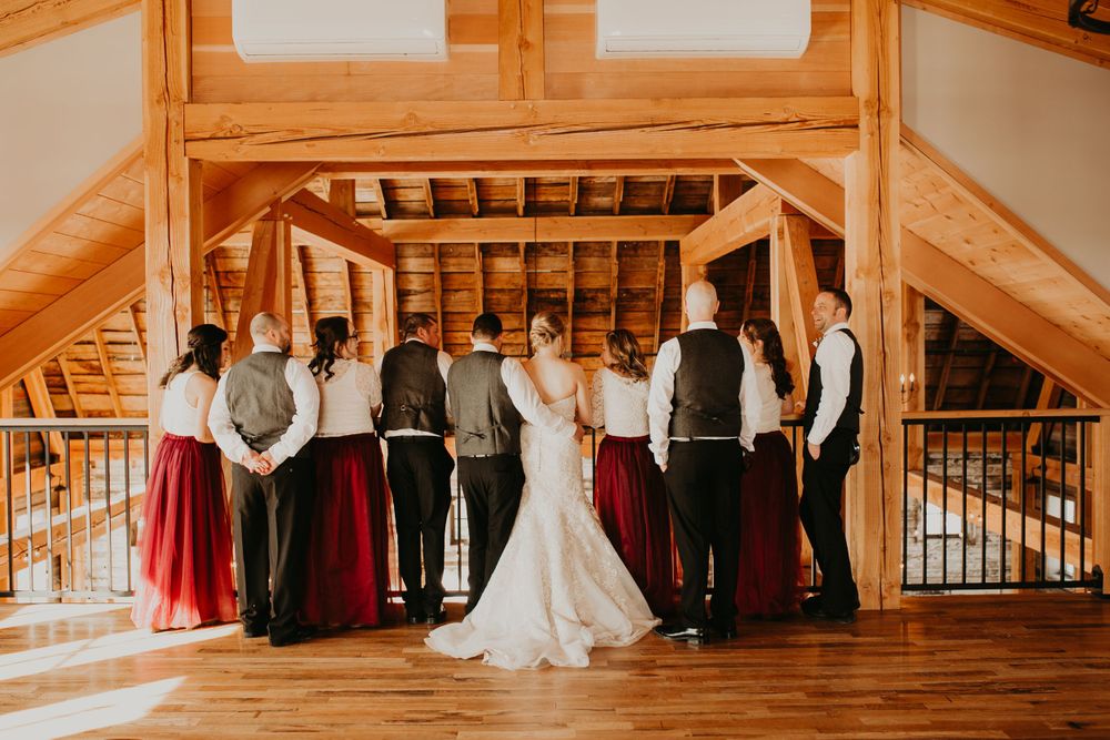 Wedding party stands with backs to camera in rustic timber-frame barn loft setting.
