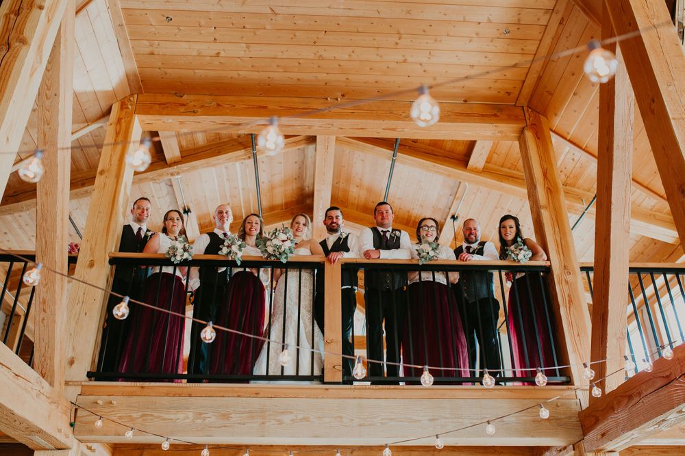 Wedding party gathers on a decorated balcony with string lights in a rustic barn venue.