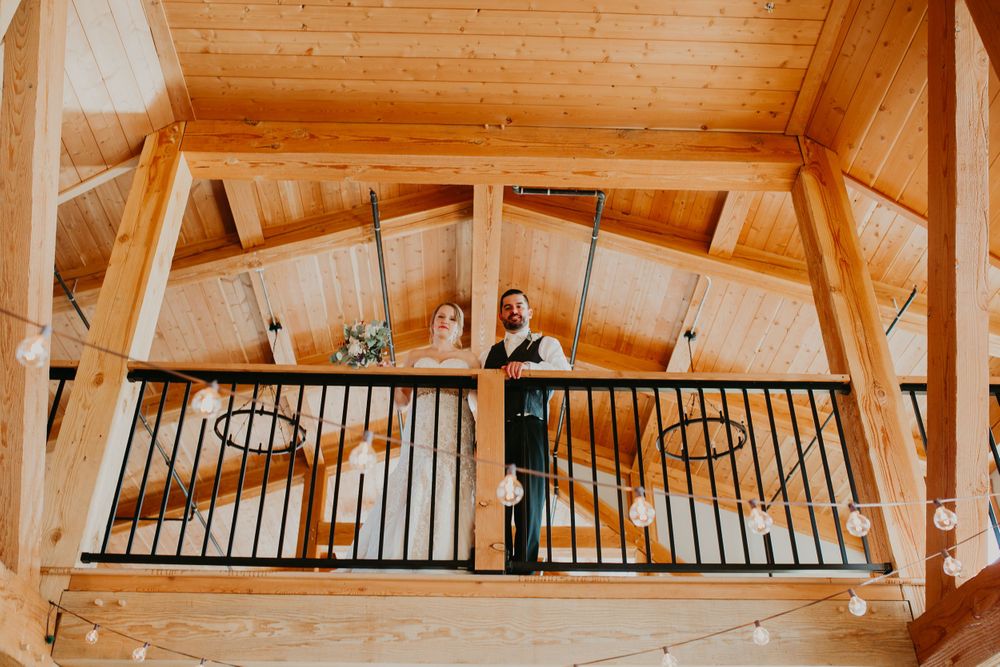 Bride and groom smile down from a wooden balcony with string lights in a barn venue.