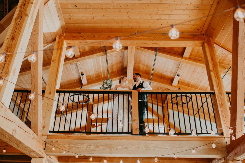 Groom stands alone on rustic barn balcony surrounded by warm wood beams and string lights.