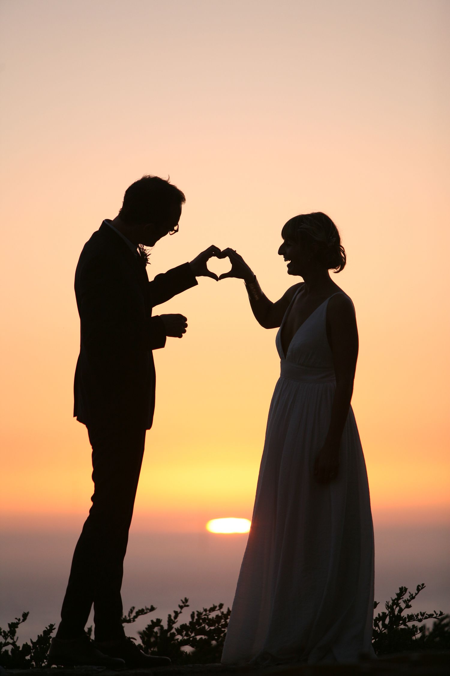 Two silhouettes make a heart shape with their hands against a golden sunset sky during a romantic moment.