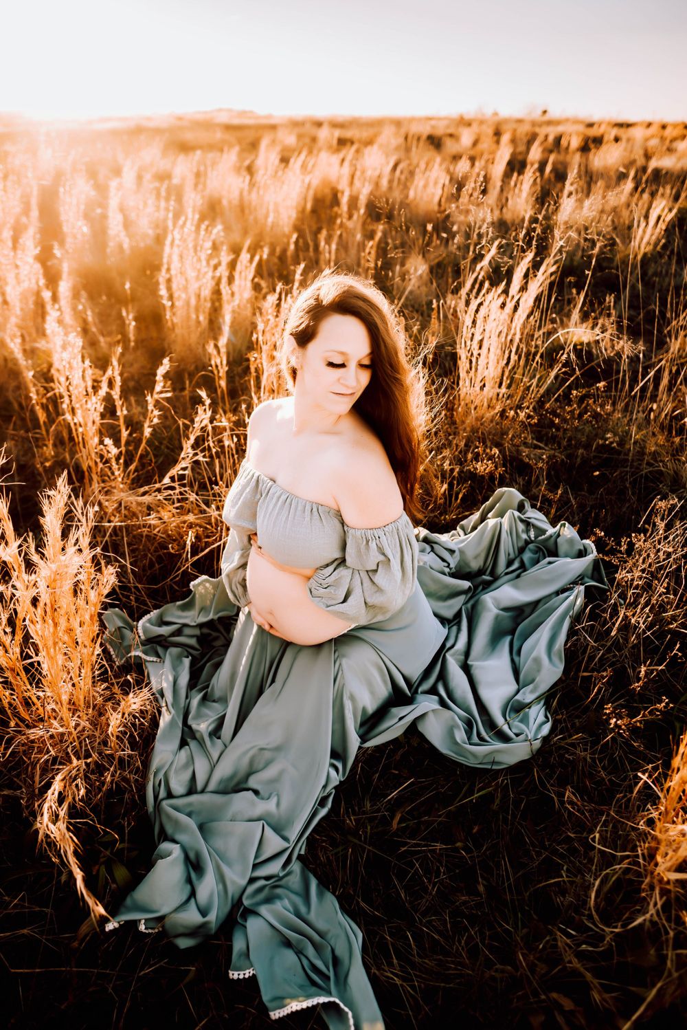 Elegant poses in a sage green dress against tall grass at sunset.