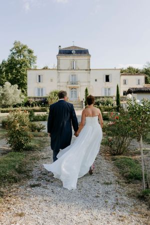 Couple walking through gravel path towards elegant French countryside mansion.