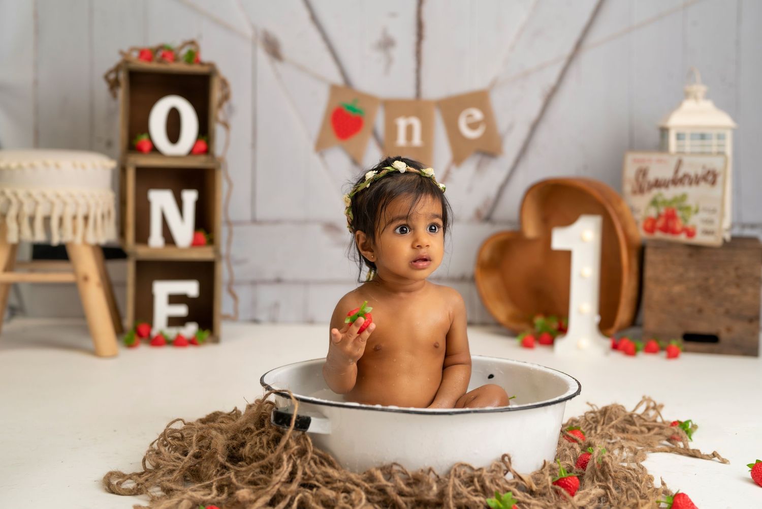Baby photoshoot with white dish and scattered red strawberries on rustic setting.