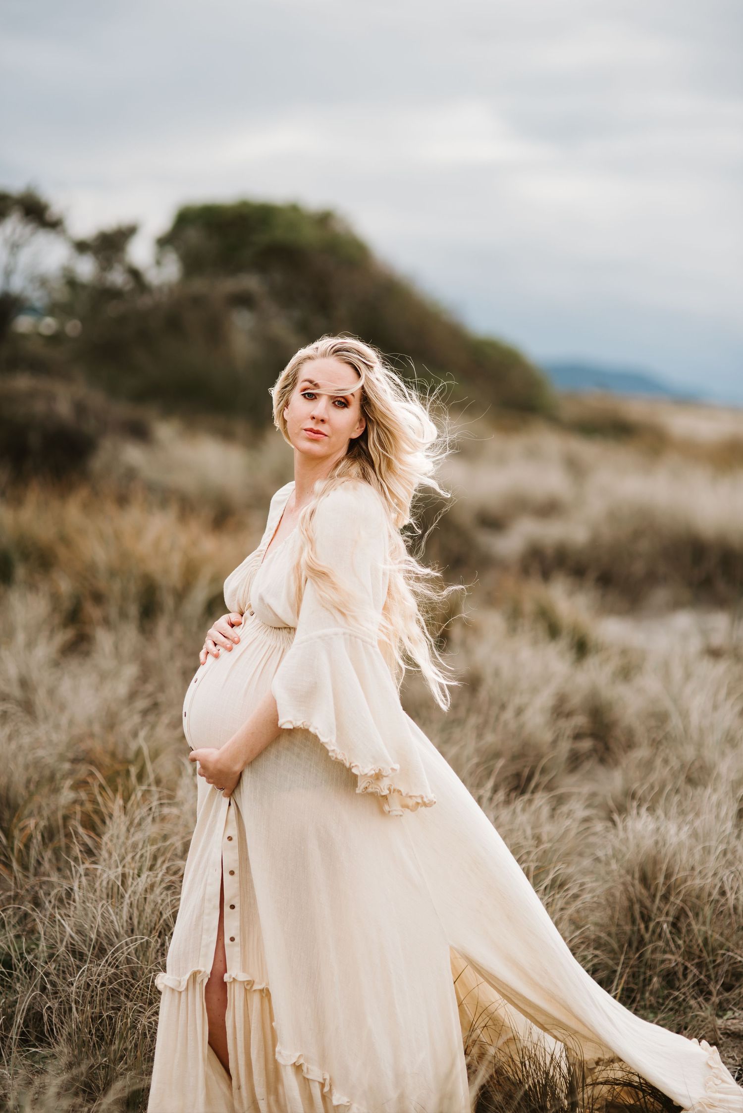 Ethereal maternity photo of woman in flowing cream dress against coastal landscape.