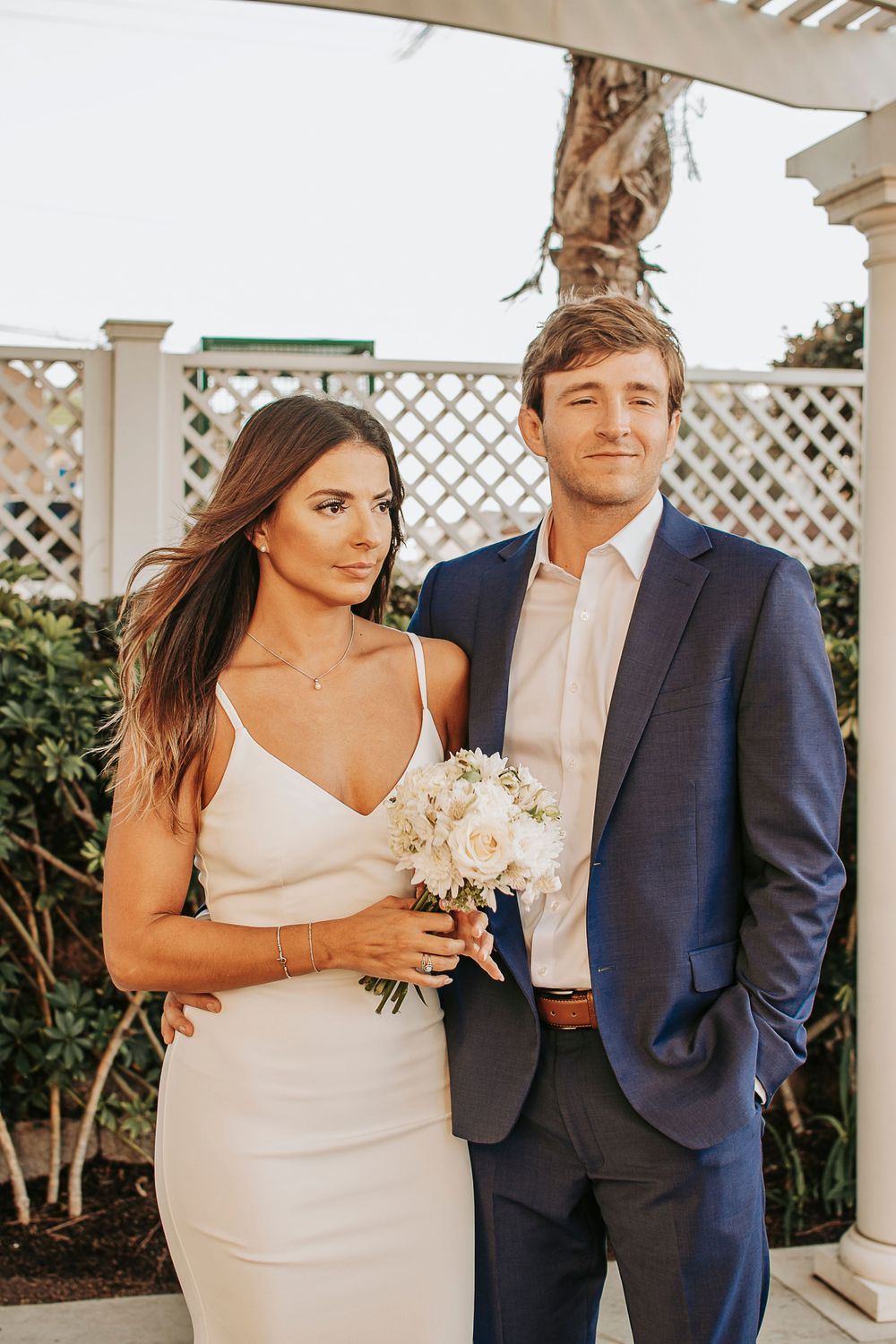 A couple in formal attire poses under a white lattice pergola with a delicate bouquet.
