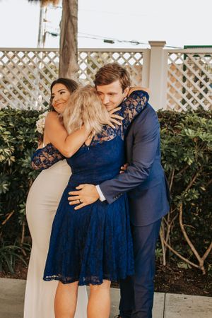 Wedding guests embrace warmly in blue and beige formal attire against a white lattice backdrop.