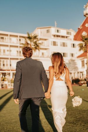 Newlyweds walk hand in hand on grass with hotel and palm trees in background.
