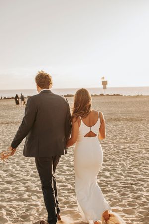 A couple walks hand in hand on a sandy beach at golden hour with a lighthouse in the distance.