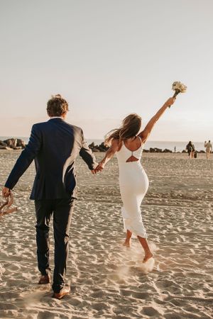 Newlyweds walking hand in hand on beach at sunset with bouquet raised in celebration.