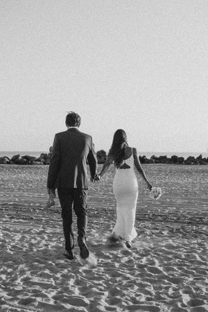 Black and white photo of couple walking along beach holding hands at sunset.