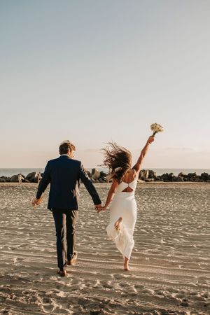 Couple runs joyfully on beach holding hands while bride raises bouquet in celebration.