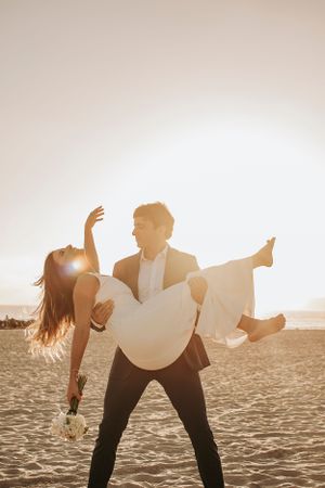 Silhouetted couple running joyfully on beach with bouquet raised against sunset sky.