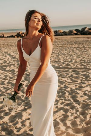 A woman in a fitted white dress poses on a beach at golden hour.