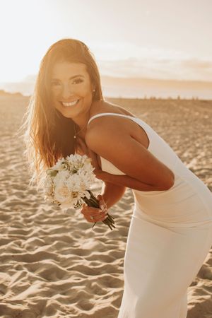 A joyful bride in a white dress poses with her bouquet during a beach photoshoot.