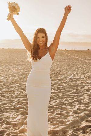 A bride celebrates on the beach with arms raised holding her bouquet at sunset.