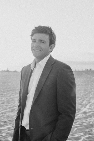 A black and white portrait of someone in a suit and tie smiling on the beach.