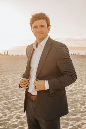 Someone wearing a dark suit adjusts their jacket while standing on a beach at sunset.