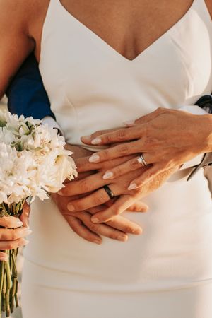 Close-up of hands with wedding rings holding a white peony bouquet.