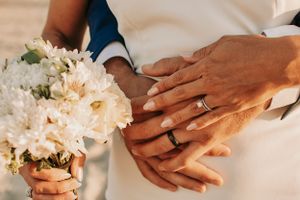 Close-up detail of newlywed hands with rings holding a white flower bouquet.