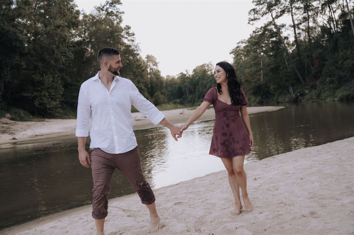 Couple playfully walking hand in hand along riverbank in casual romantic photoshoot.