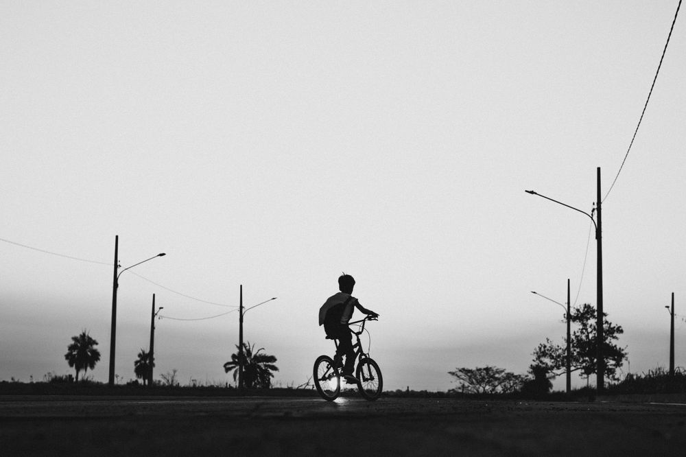 Black and white silhouette of cyclist riding along road with street lamps.