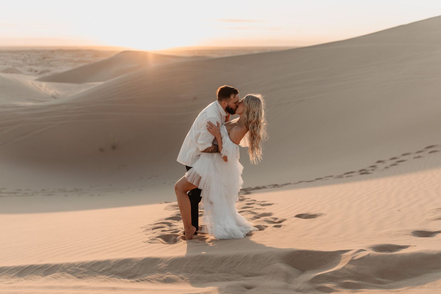 A romantic desert engagement photo with footprints in sand dunes at sunset.