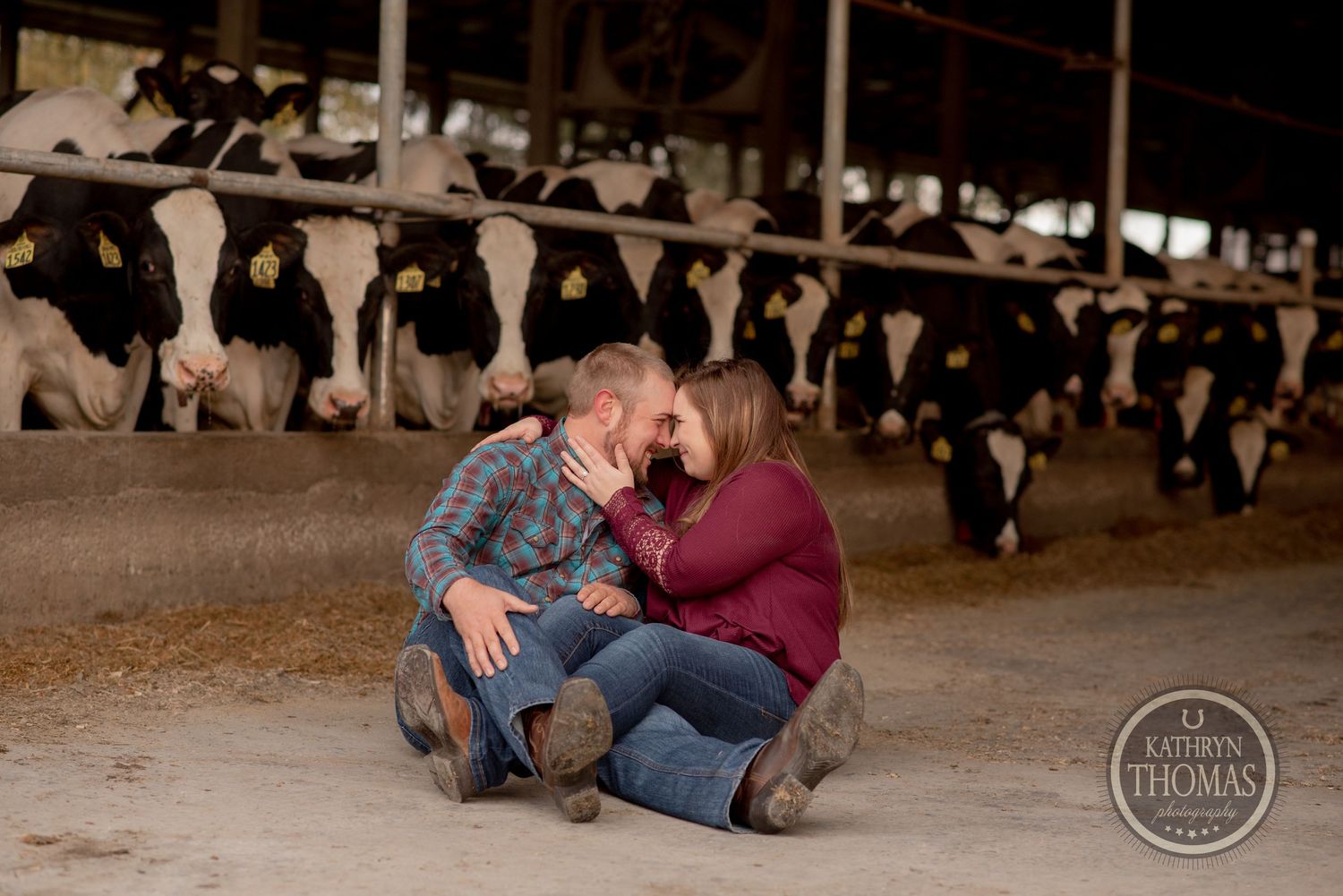 Couple sits together on barn floor with Holstein dairy cows in background.