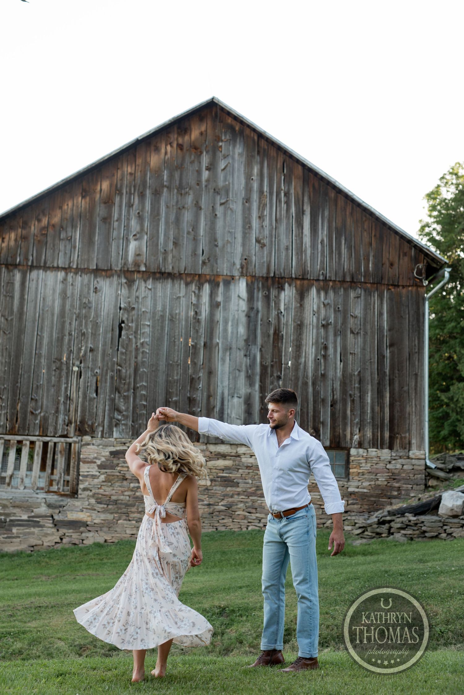 A couple dances playfully in front of a rustic wooden barn during a summer photoshoot.