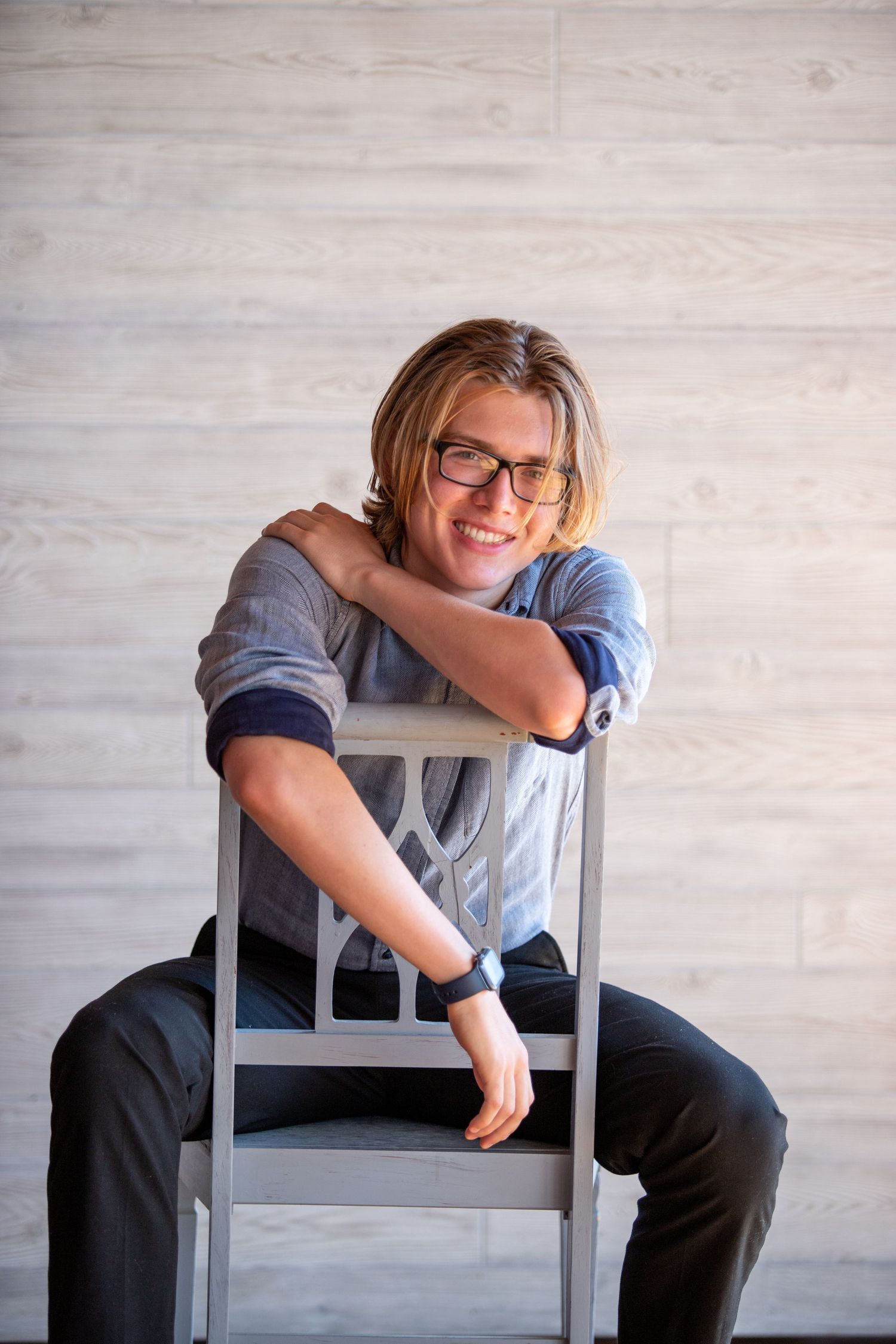 Smiling person with glasses leaning on a gray chair against a light wood background.