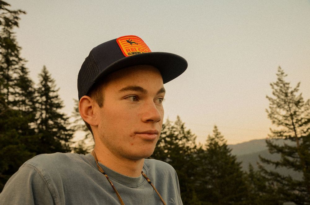 Person in baseball cap with mountains and evergreen trees in background during golden hour.