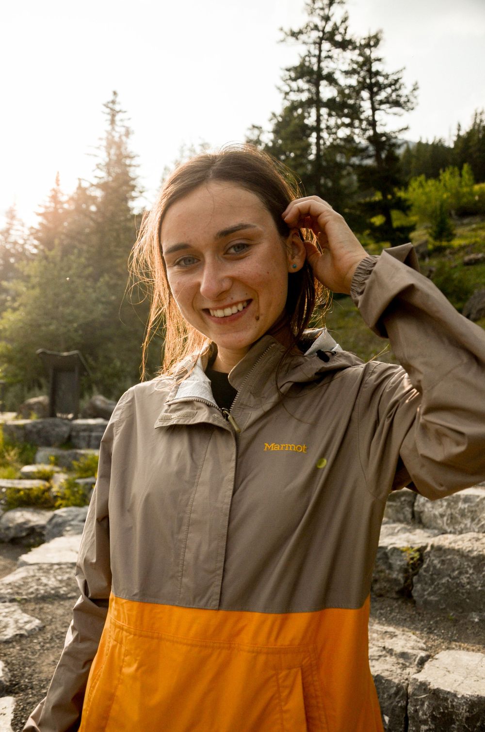 Person in tan and orange jacket smiling outdoors with sunlit forest background.