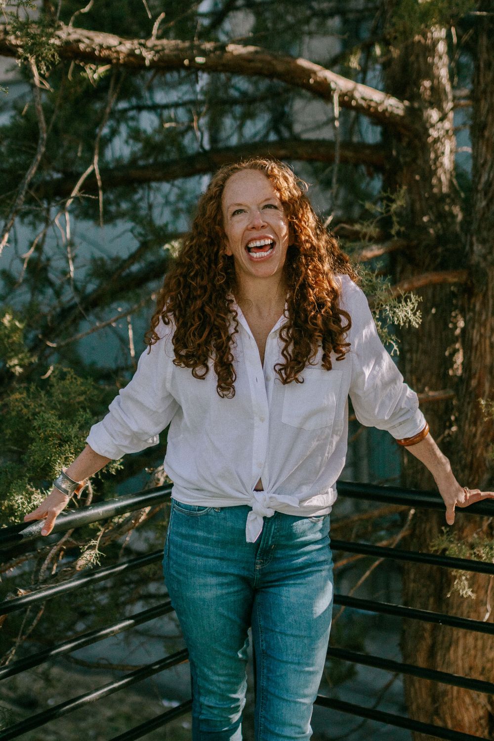 A joyful person with curly hair wearing a white tied shirt and jeans laughs while standing in a wooded setting.