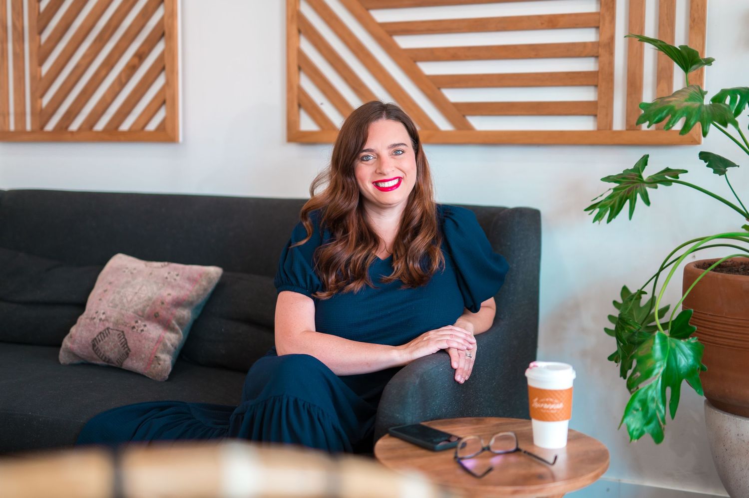 Smiling woman wearing a blue dress sitting on a blue couch looking comfortable with glasses and coffee nearby; boho vibe