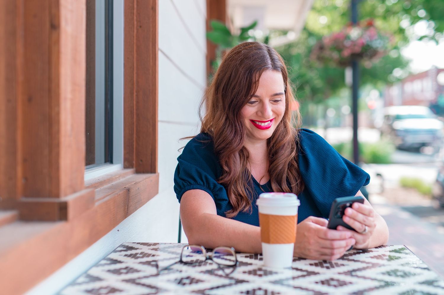 Woman working on her phone with glasses and coffee on table; set outside