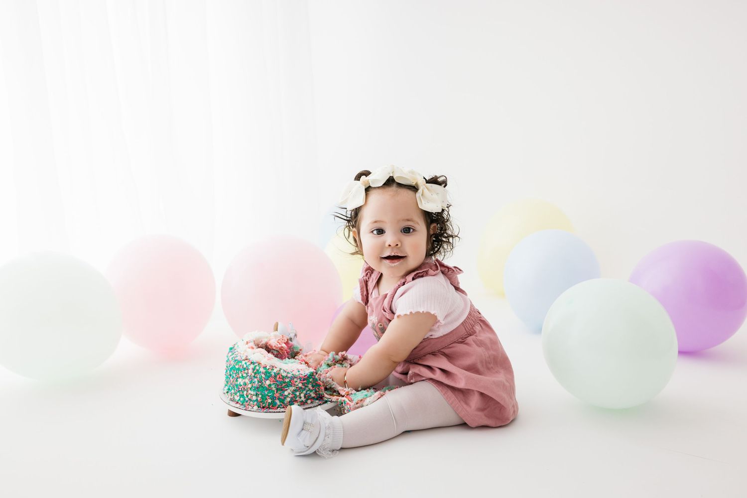 Little girl with pigtails and bows smashing her hands into cake during her cake smash 1st birthday photography session.