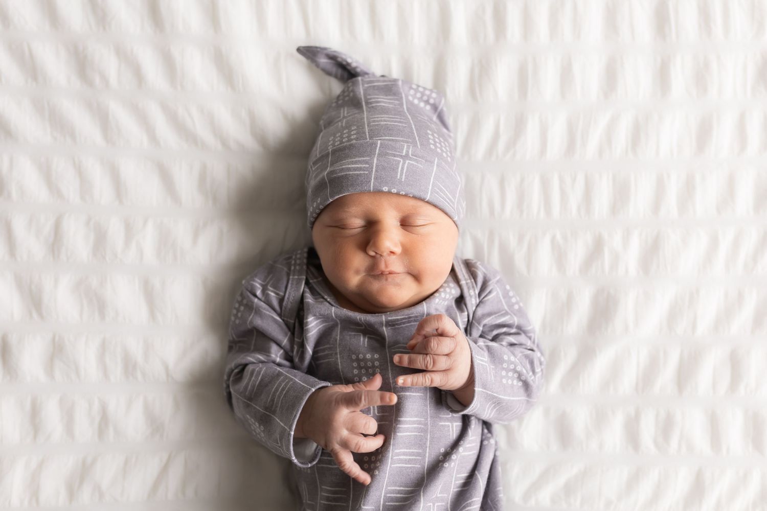 Baby wearing grey outfit and hat lying on textured white background.