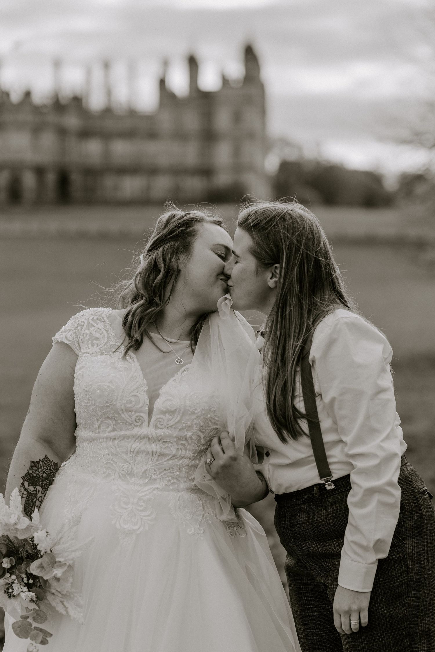 a black and white image of two brides kissing, standing in from of a stately home.