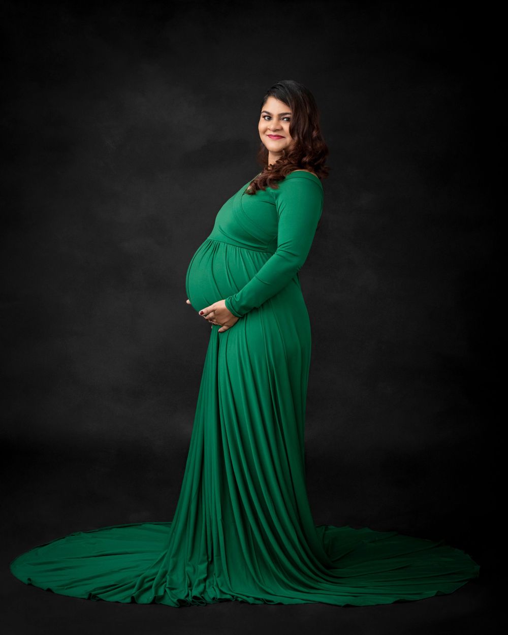 Elegant maternity portrait in emerald green flowing gown against dark backdrop.