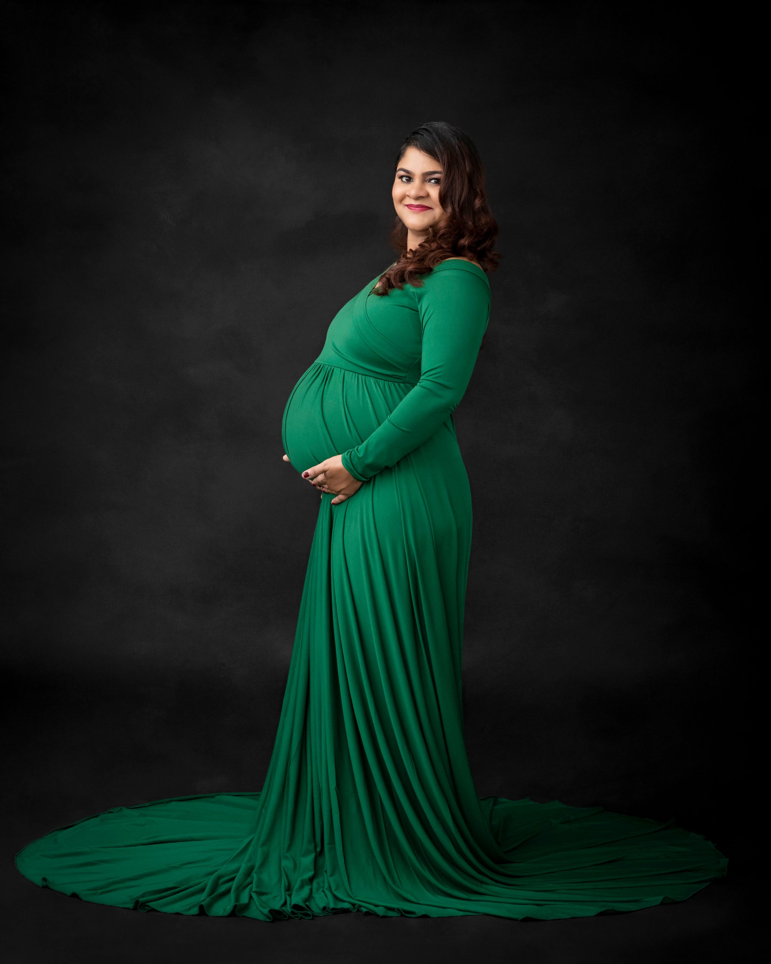 Elegant maternity portrait in emerald green flowing gown against dark backdrop.