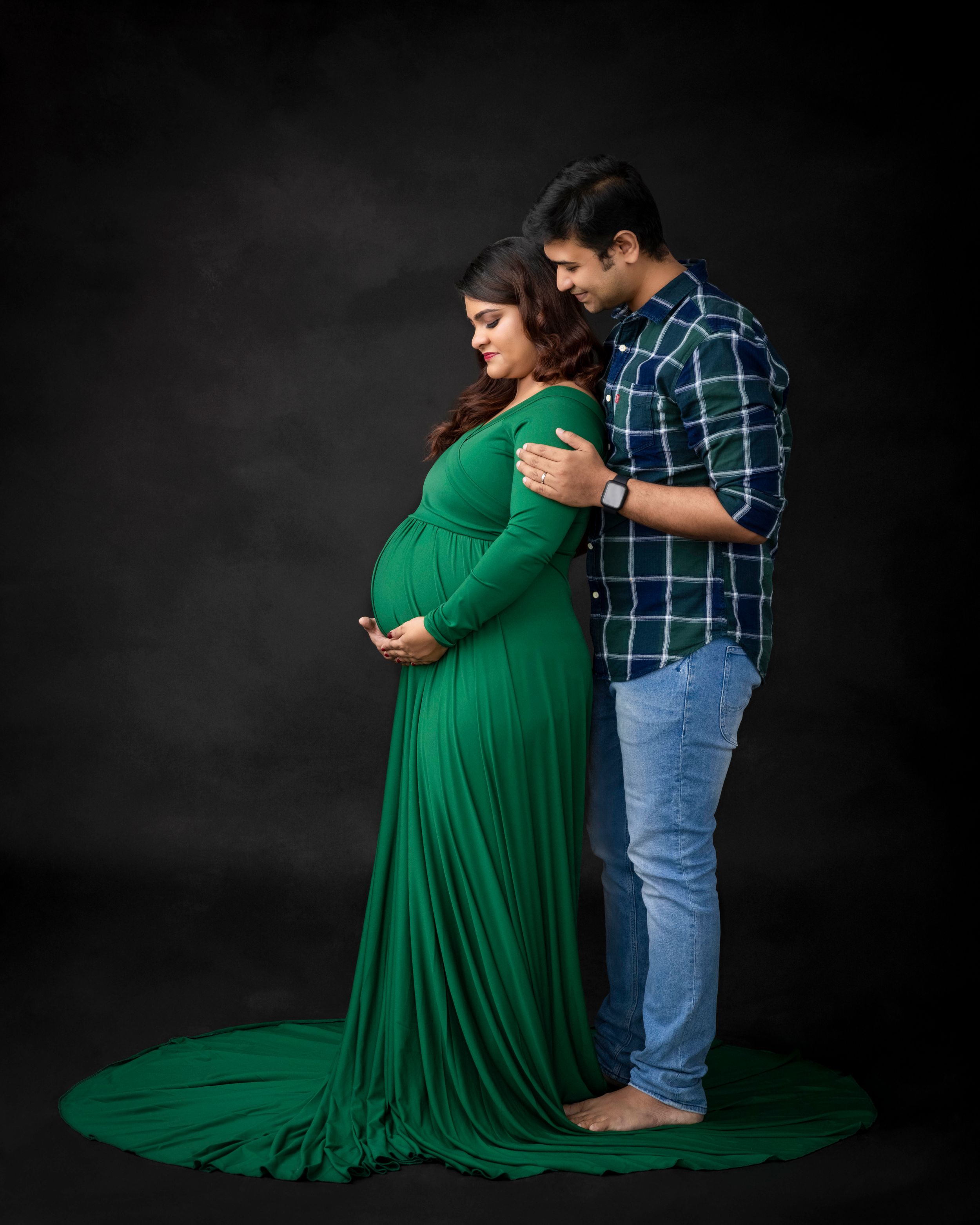 Intimate maternity portrait with couple in green dress and plaid shirt against dark background.