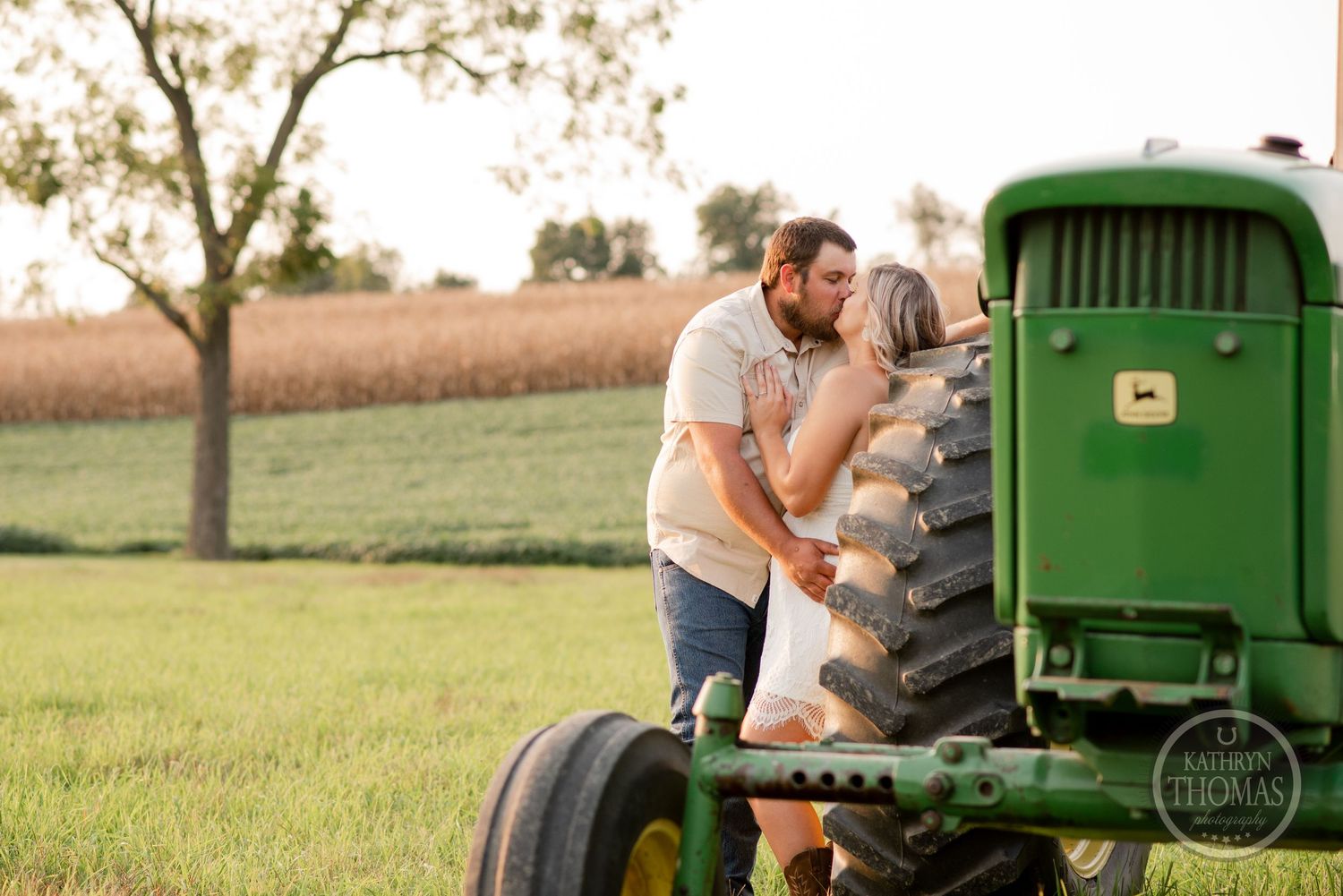Couple embracing on the large wheel of an antique green tractor in a sunlit farm field.