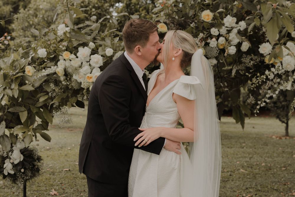 Romantic wedding couple sharing intimate moment under white floral arch in garden setting.