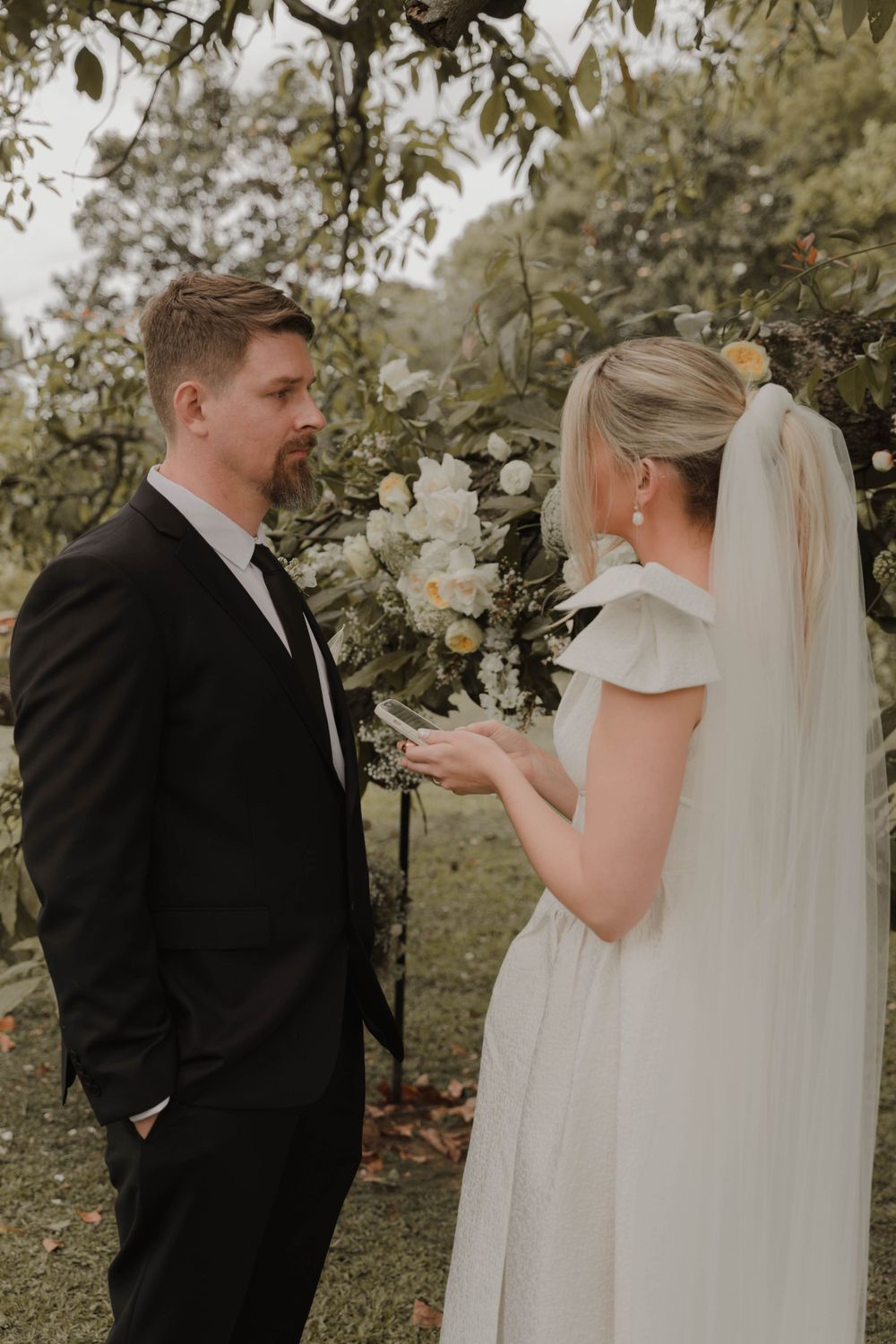 A bride in white dress and veil speaks to her groom under a floral garden arch.