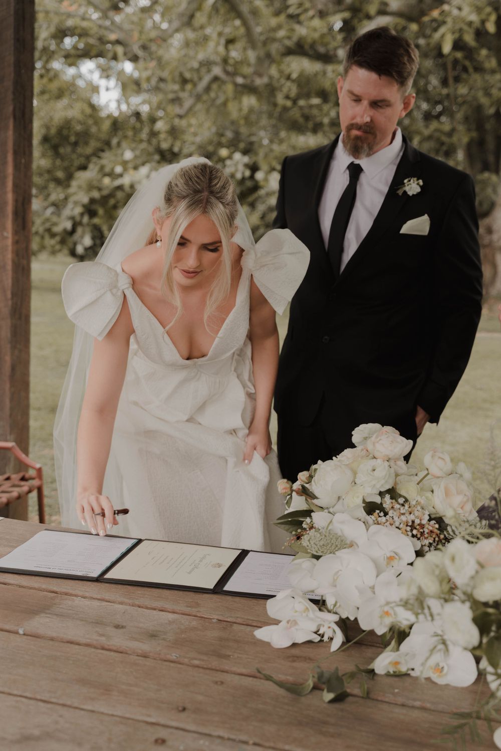 Couple signing marriage documents at rustic wooden table decorated with white flowers.