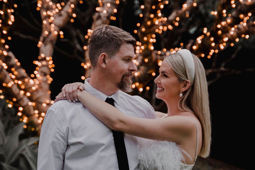 Joyful embrace under trees decorated with warm glowing string lights at dusk.